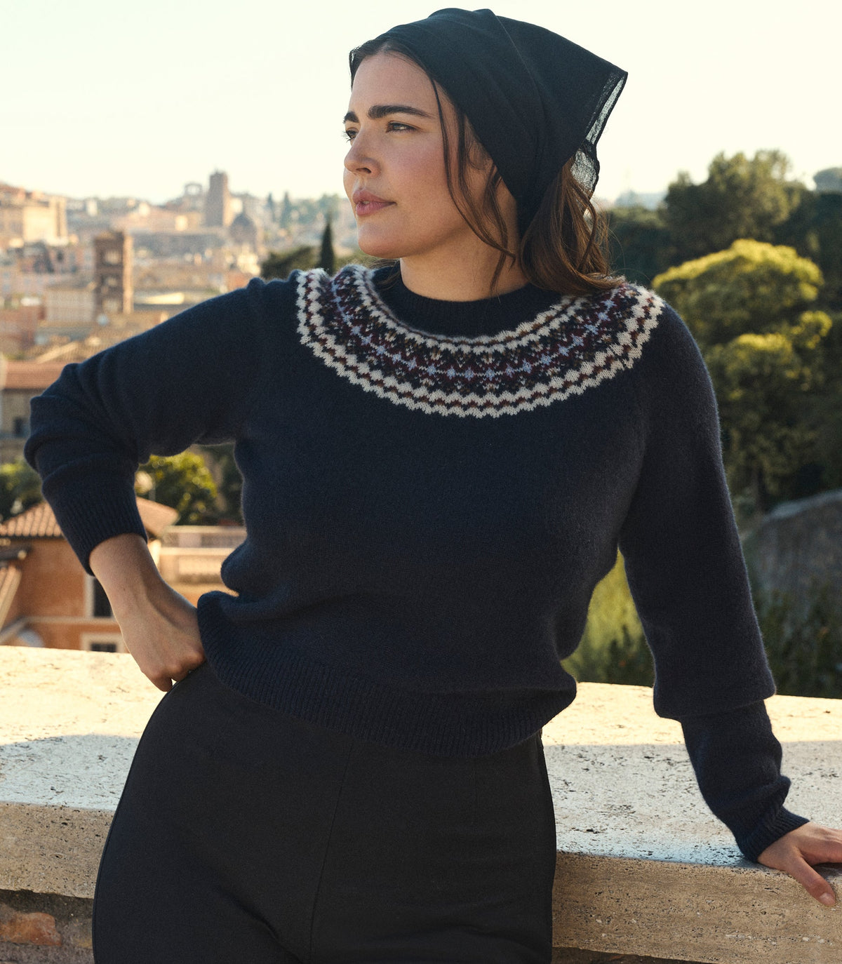 A woman wearing a dark navy knit sweater with a white and purple Fair Isle yoke pattern, paired with a black bandana, posing outdoors against a scenic city background.