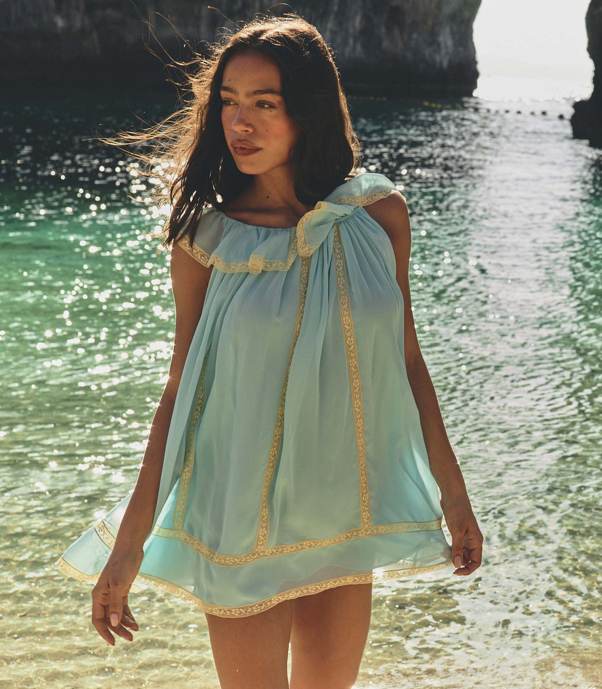 Woman in a light blue dress standing by a body of water with sunlight filtering through.
