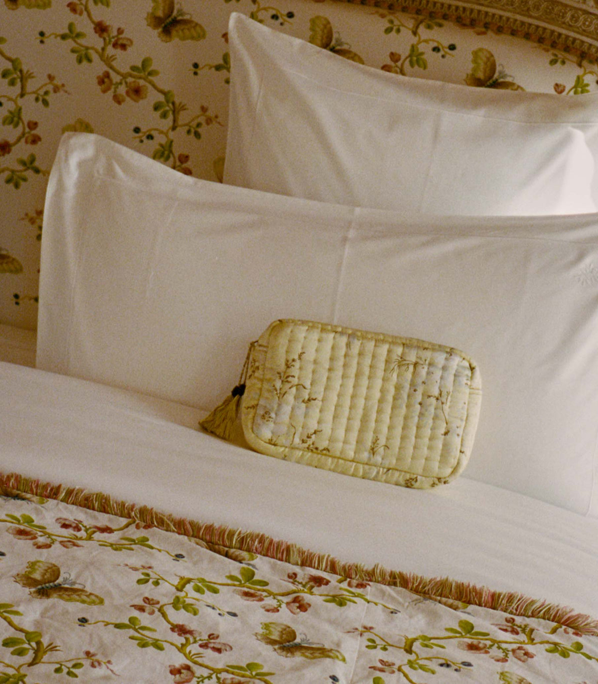 Light yellow floral pouch against white pillows on a bed with floral bedding and wallpaper.