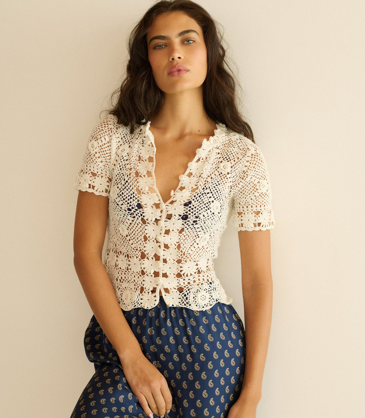Woman wearing a white crochet top and blue patterned pants against a beige background