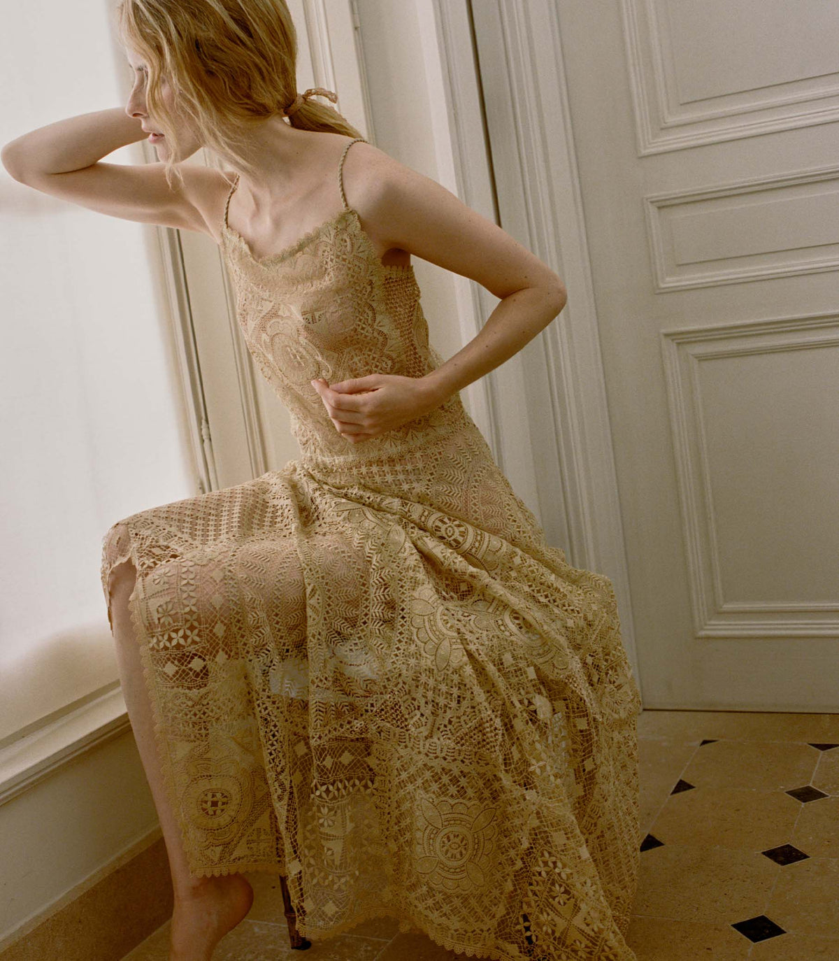 Woman wearing a pale gold crochet dress in an elegant room with white walls and a patterned floor.